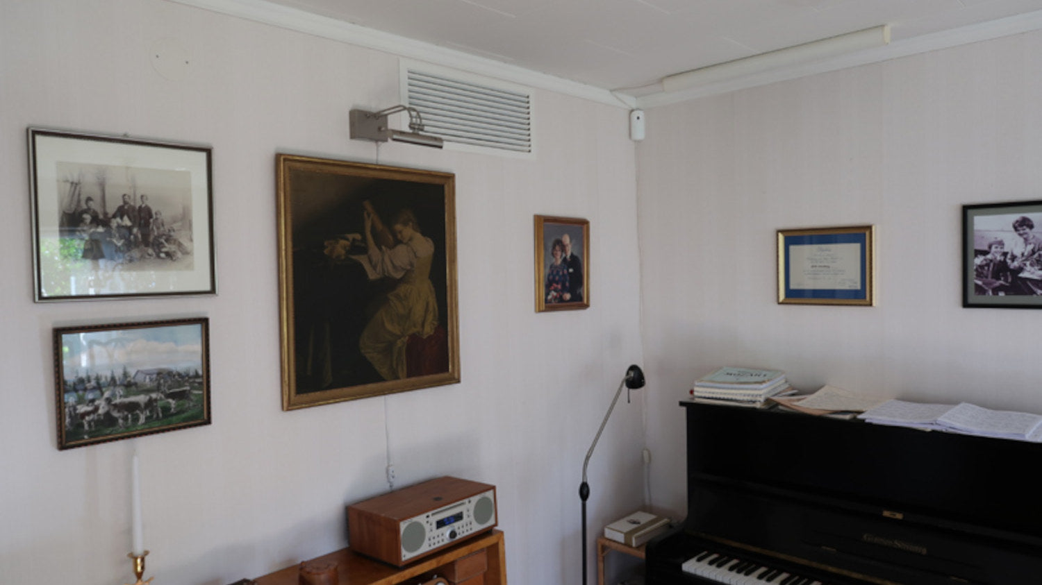 Room interior with framed pictures on the wall, a piano, and a radio and Mobair Room 2 Room heat transfer fan installed inside of the wall near the ceiling.